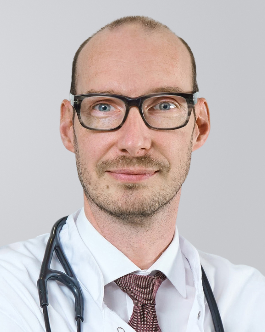 Ein Arzt mit Brille, kurzen Haaren und weißem Kittell vor einem neutralen Hintergrund.