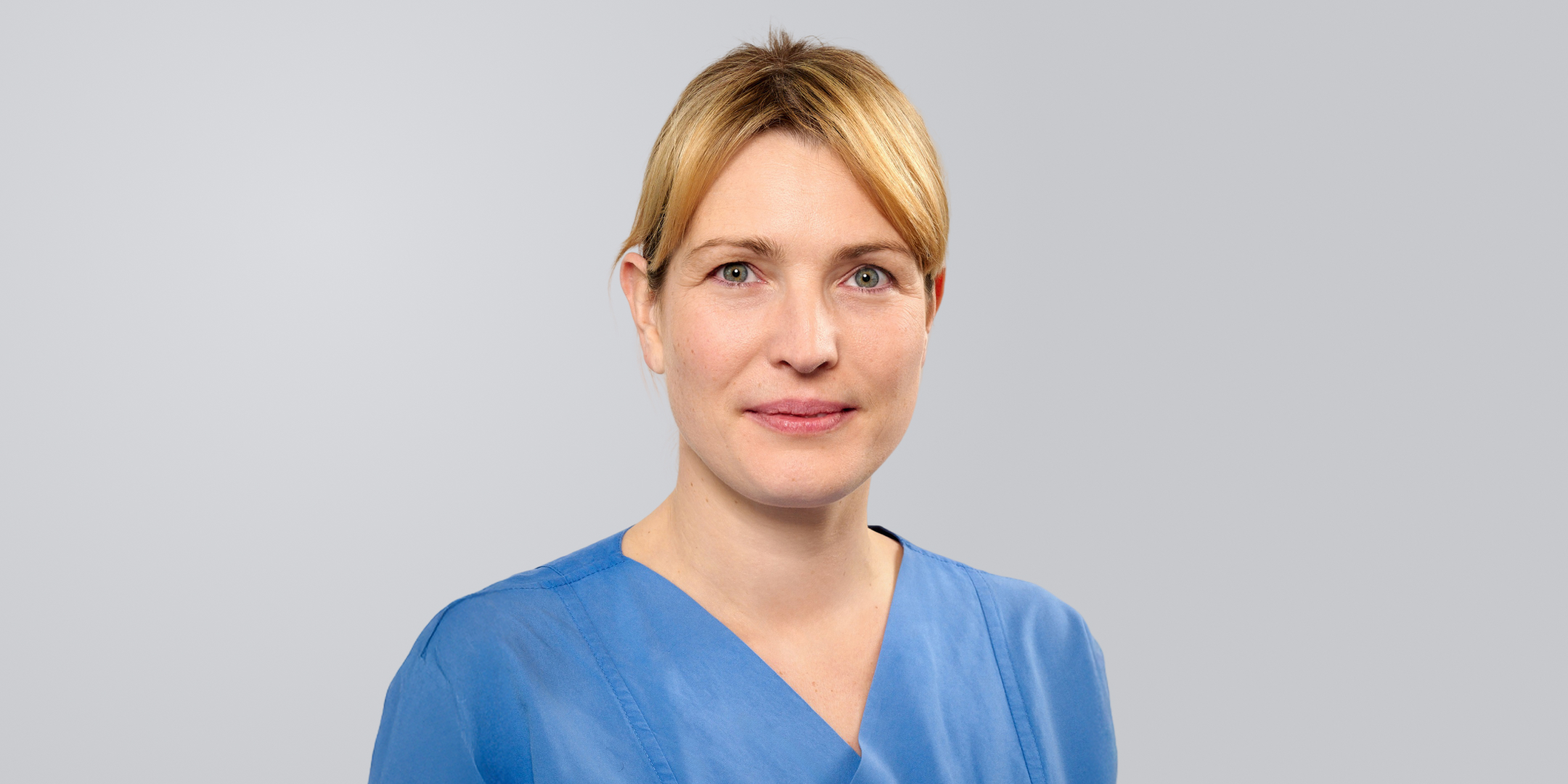 Frau mit blonden Haaren in blauer Arbeitskleidung vor einem grauen Hintergrund.