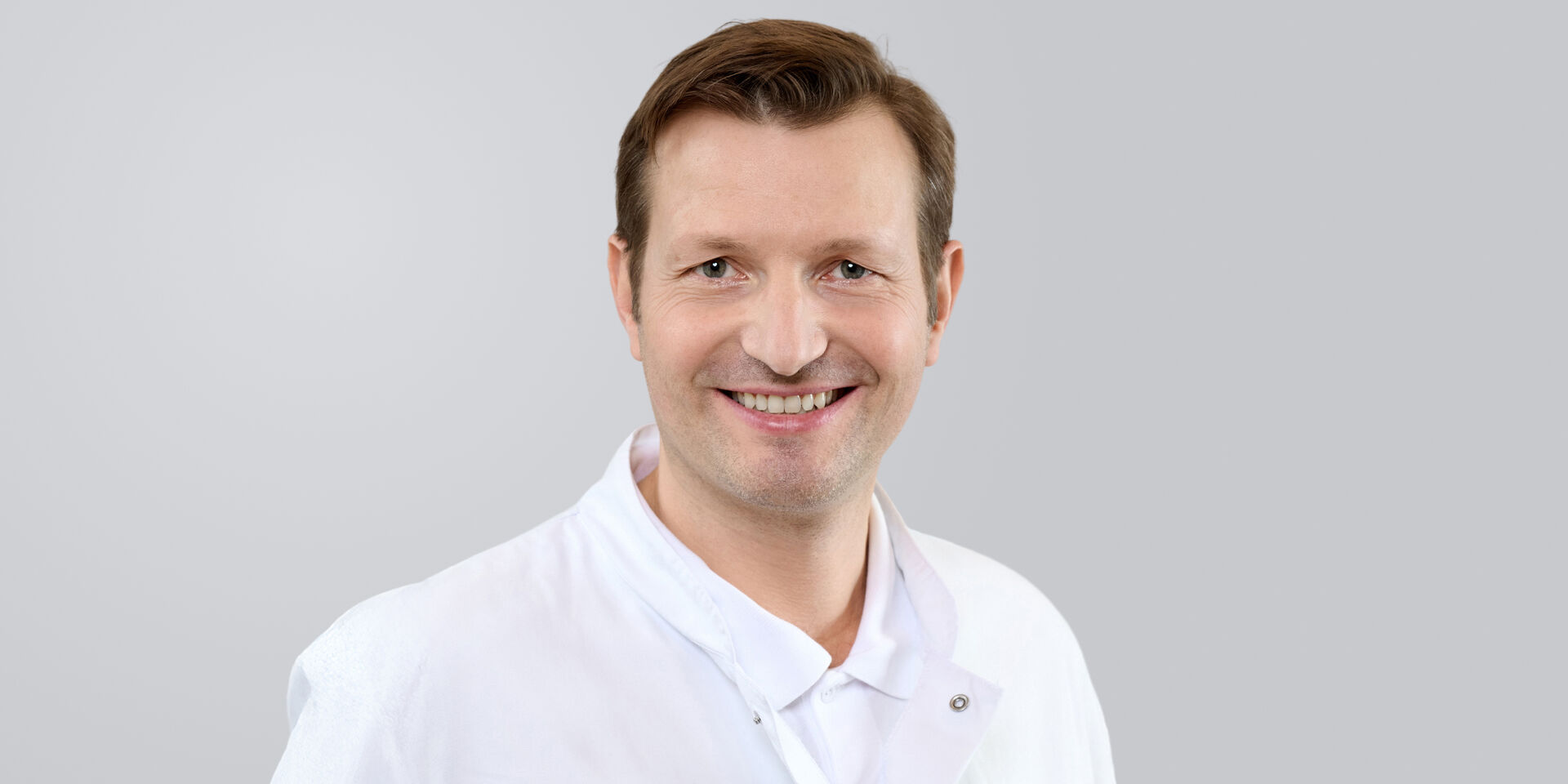 Lächelnder Mann in weißem Arztkittel vor grauem Hintergrund.