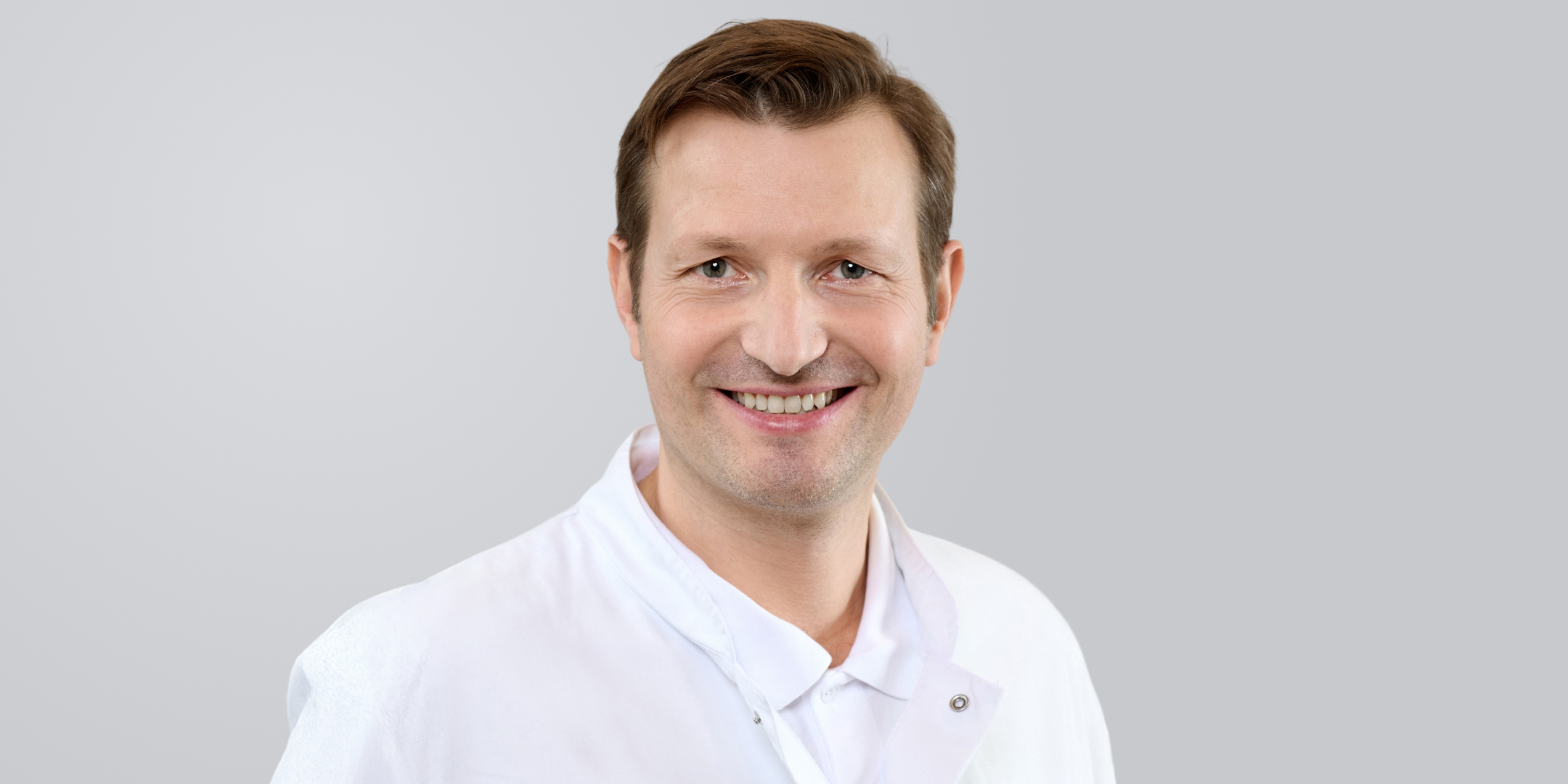Lächelnder Mann in weißem Arztkittel vor grauem Hintergrund.