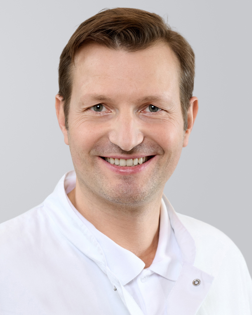 Lächelnder Mann in weißem Arztkittel vor grauem Hintergrund.