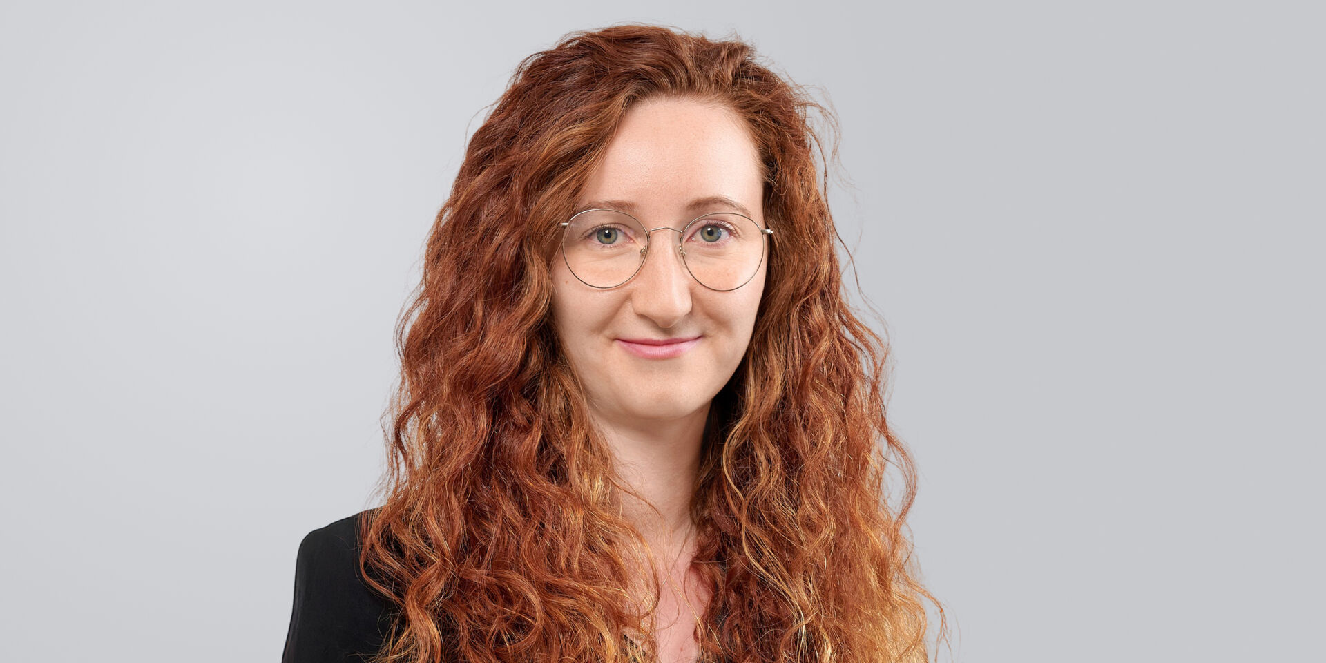 Frau mit lockigem, rotem Haar und Brille lächelt vor einem grauen Hintergrund.