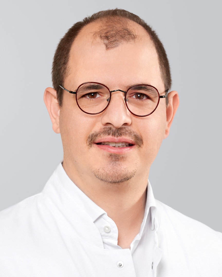 Porträt eines Mannes mit Brille, kurzem Haar und weißem Kittel vor neutralem Hintergrund.