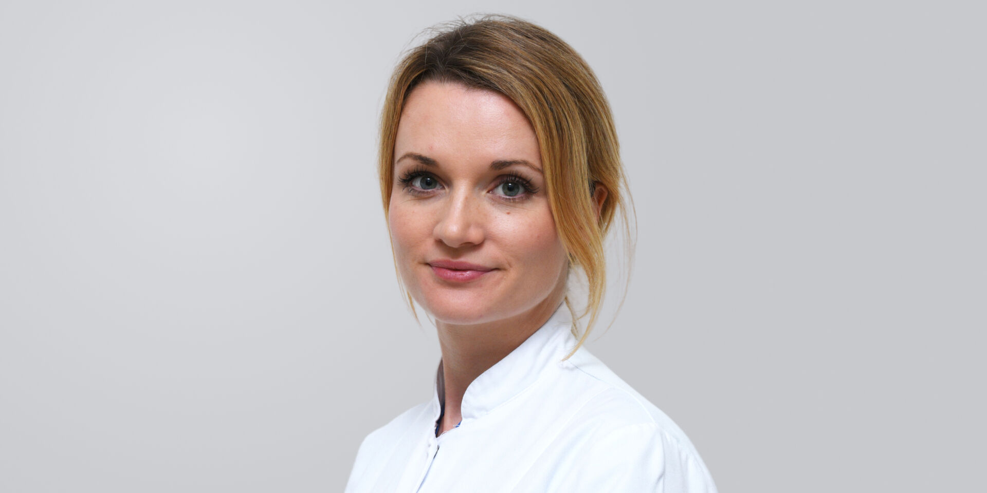 Junge Frau mit blonden Haaren in einem weißen Kittel vor einem grauen Hintergrund.