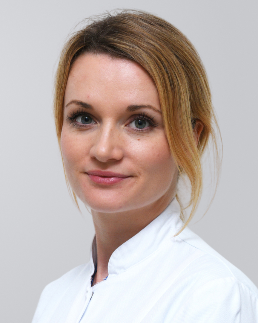 Junge Frau mit blonden Haaren in einem weißen Kittel vor einem grauen Hintergrund.