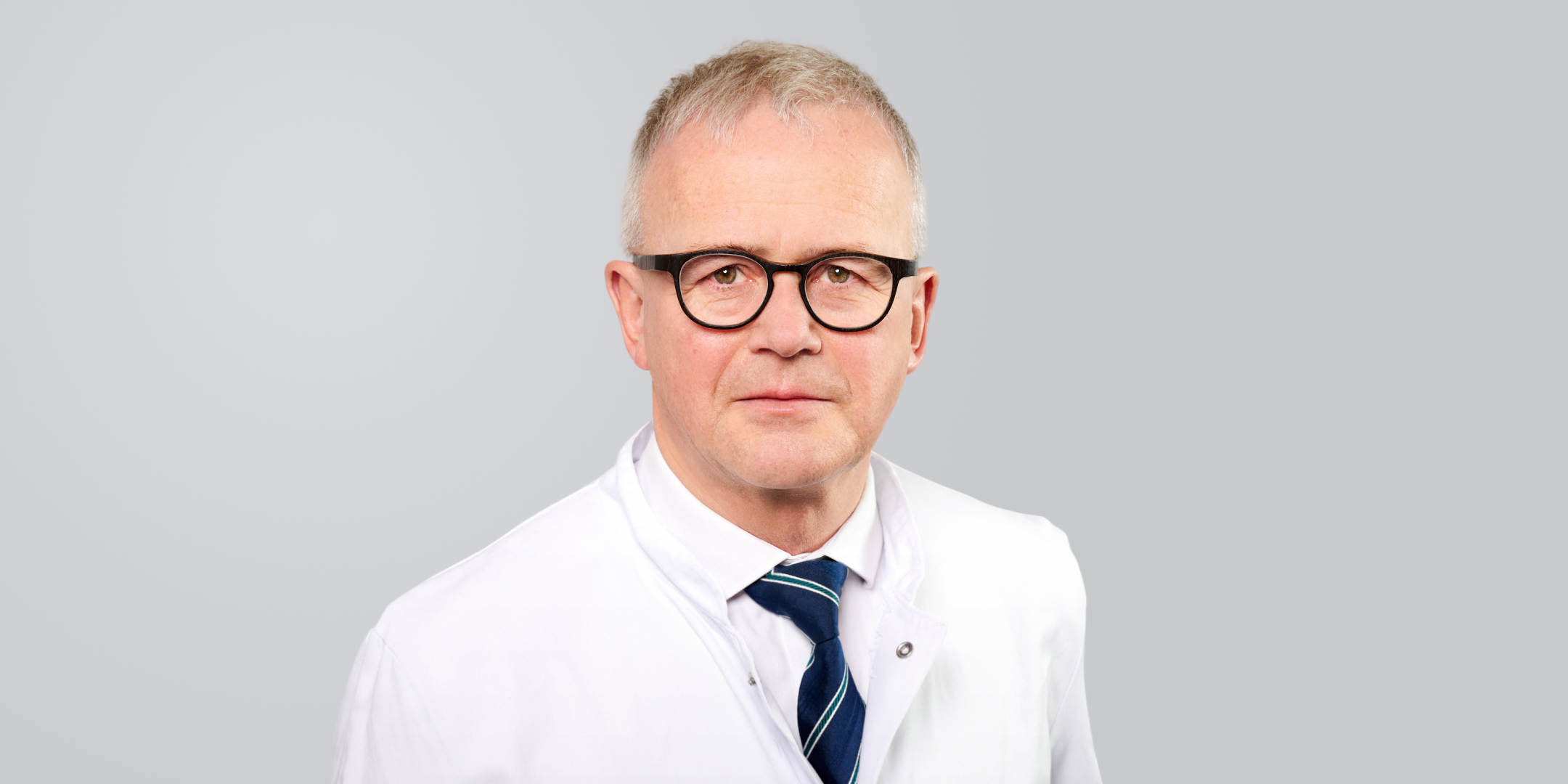 Mann mit einem weißen Arztkittel, Brille und Krawatte vor einem grauen Hintergrund.