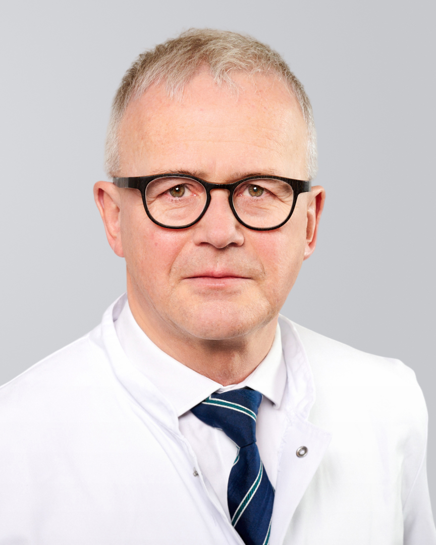 Mann mit einem weißen Arztkittel, Brille und Krawatte vor einem grauen Hintergrund.
