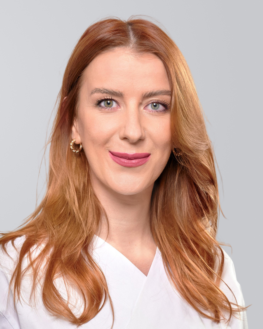 Frau mit langen, roten Haaren, weißem Kittel und einem freundlichen Lächeln vor grauem Hintergrund.