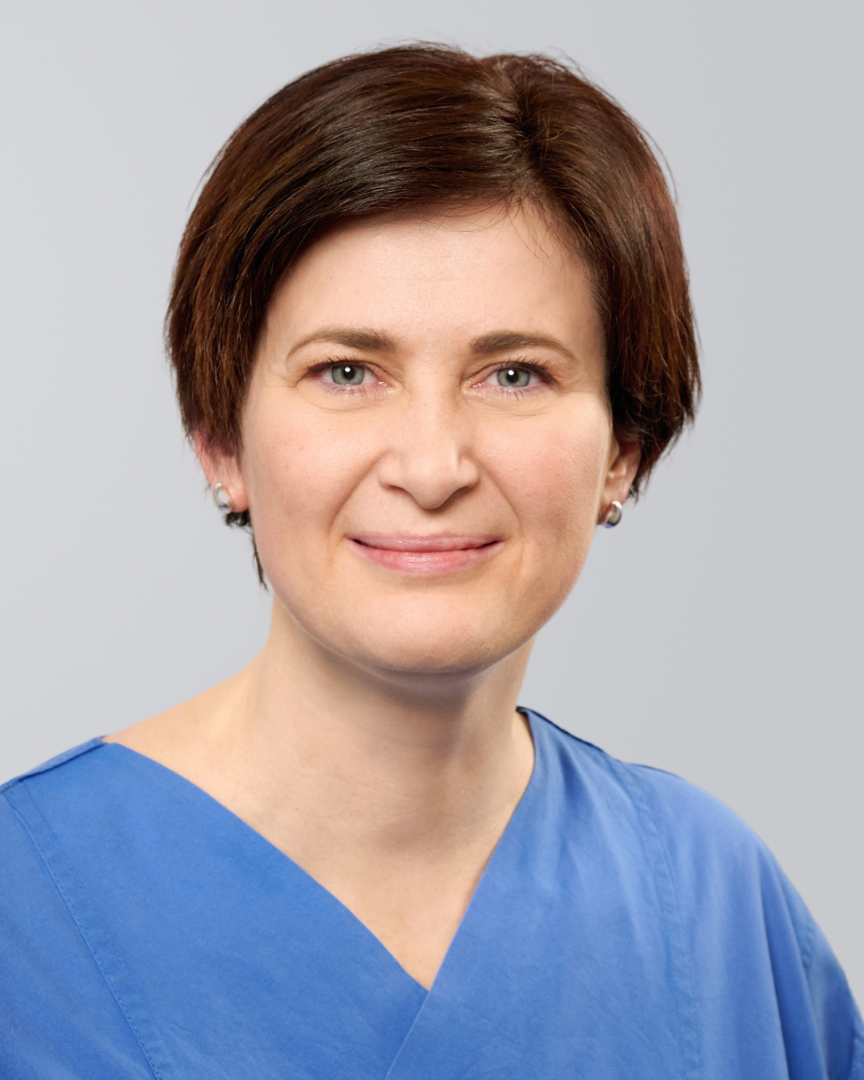 Frau in blauer medizinischer Kleidung mit kurzem, braunem Haar und freundlichem Lächeln vor neutralem Hintergrund.