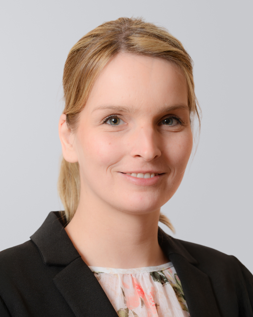 Porträt einer lächelnden Frau mit blonden Haaren und schwarzem Blazer vor einem hellen Hintergrund.