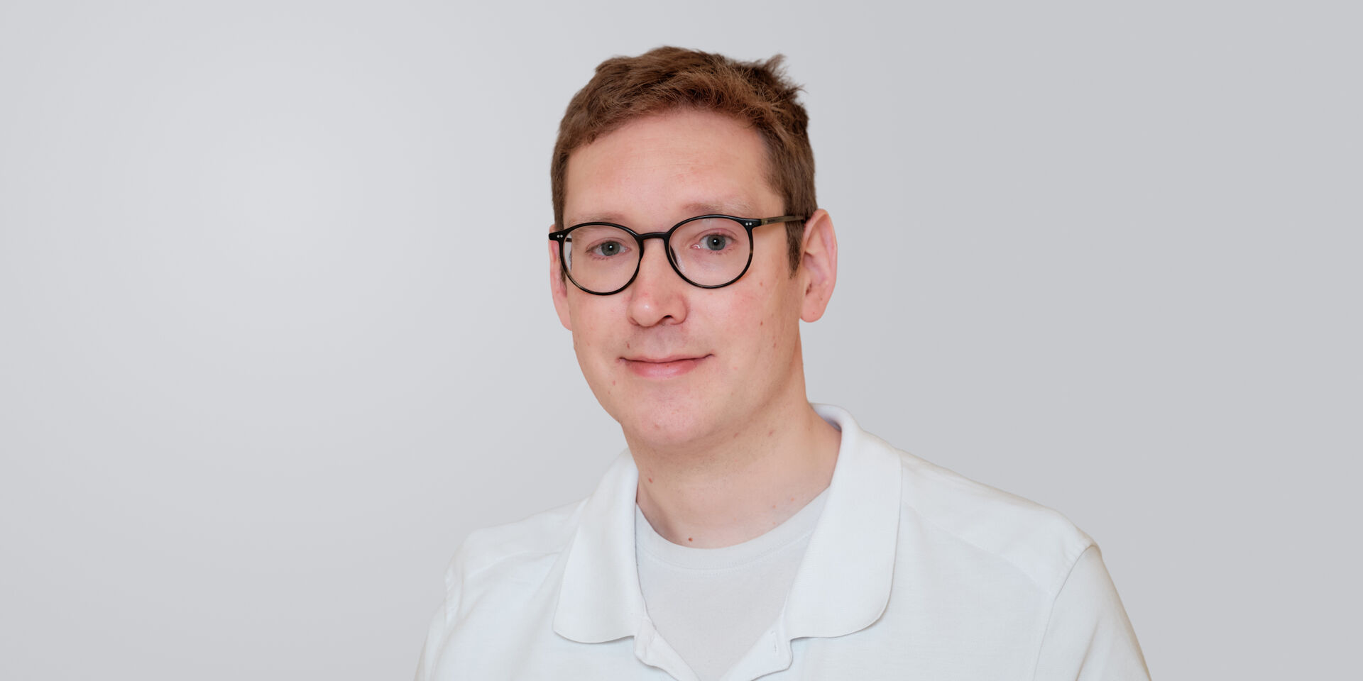 Junger Mann mit Brille und weißem Arztkittel vor einem neutralen Hintergrund.