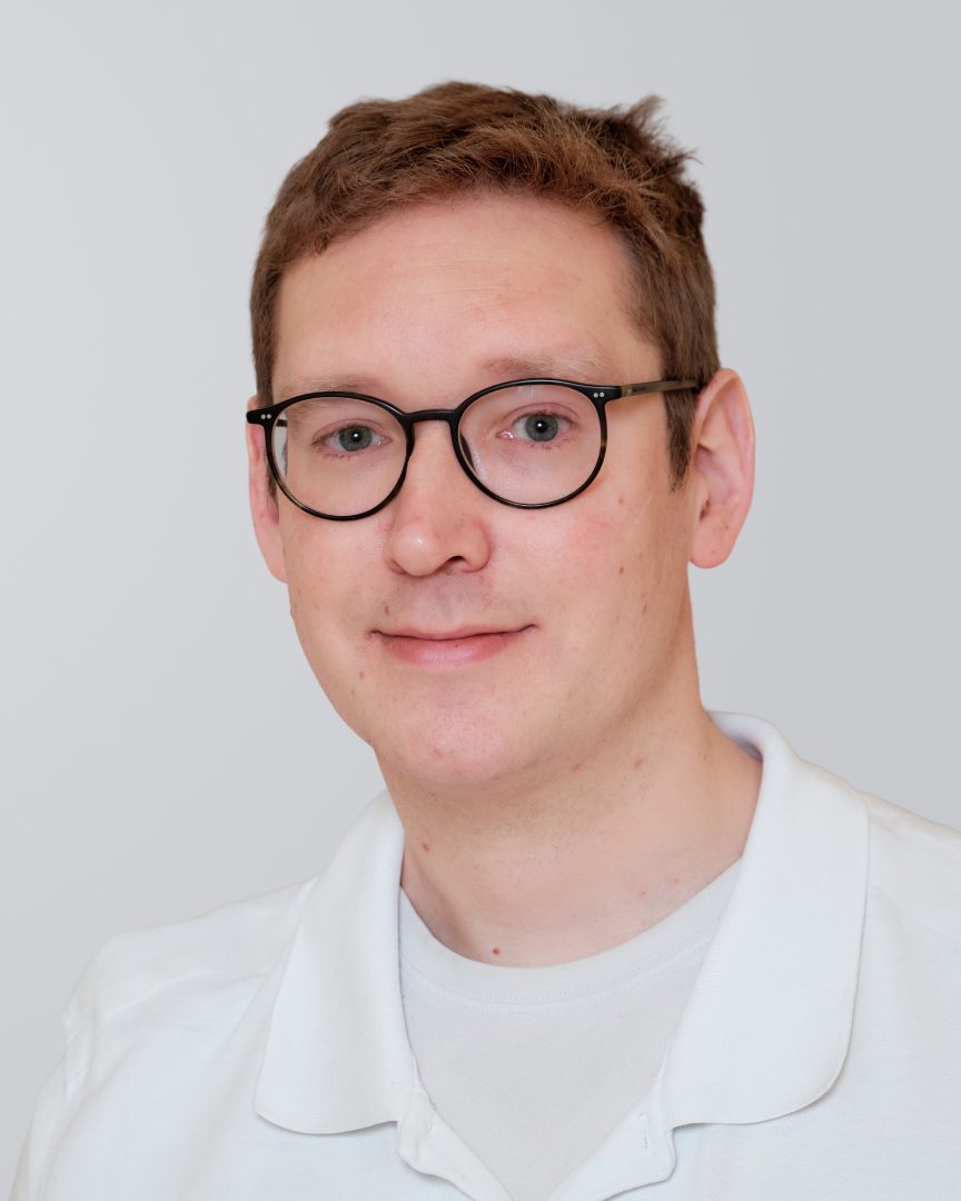 Junger Mann mit Brille und weißem Arztkittel vor einem neutralen Hintergrund.