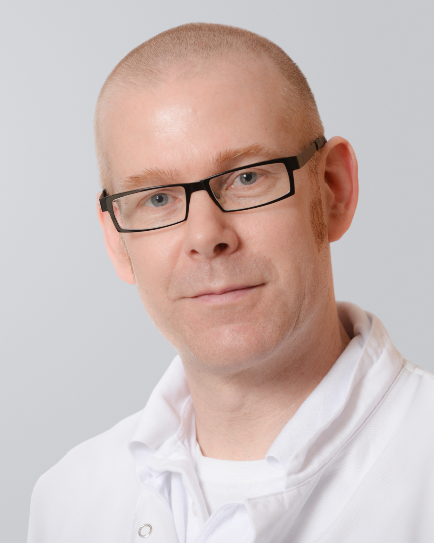 Mann mit Glatze, Brille und weißem Kittel vor einem grauen Hintergrund.