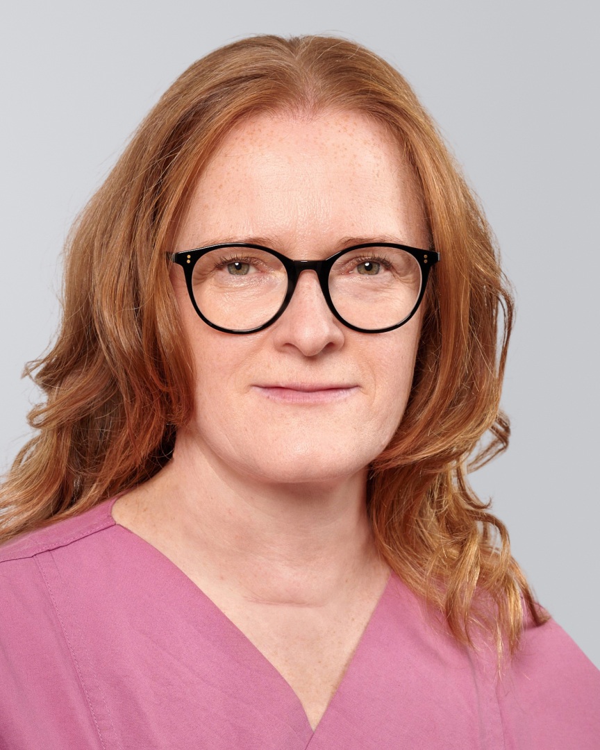 Frau mit langen, roten Haaren, Brille und pinkfarbenem Oberteil vor einem grauen Hintergrund.