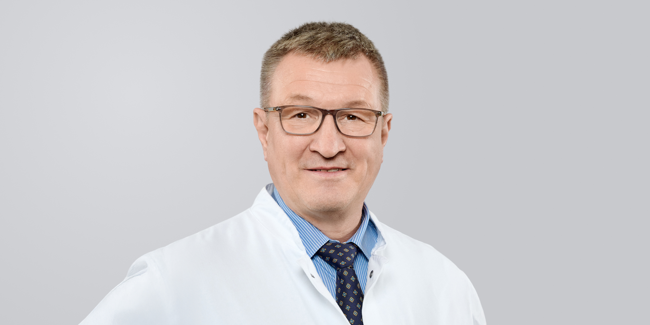 Älterer Mann mit weißem Kittel, Brille und Krawatte vor einem grauen Hintergrund.