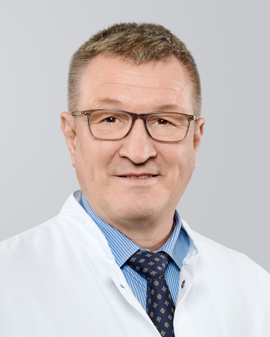 Älterer Mann mit weißem Kittel, Brille und Krawatte vor einem grauen Hintergrund.
