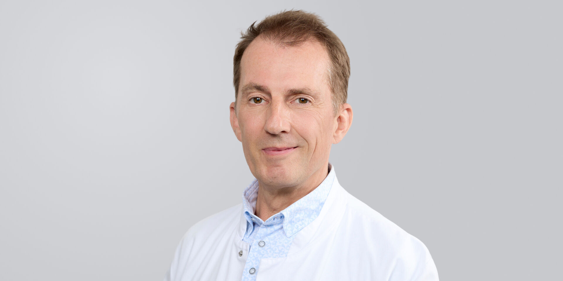 Lächelnder Mann in weißem Kittel vor neutralem Hintergrund.