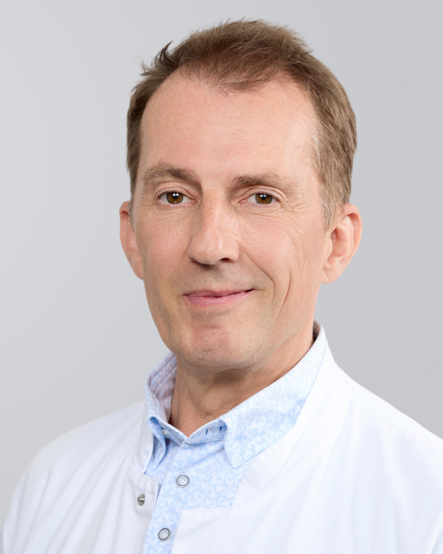 Lächelnder Mann in weißem Kittel vor neutralem Hintergrund.