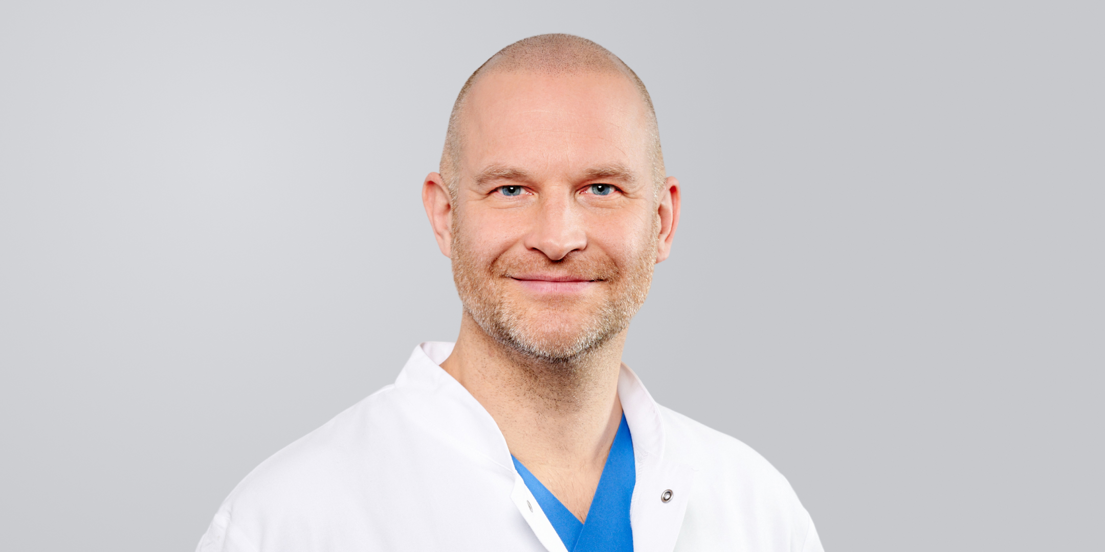 Lächelnder Mann mit Glatze und weißem Kittel vor grauem Hintergrund.