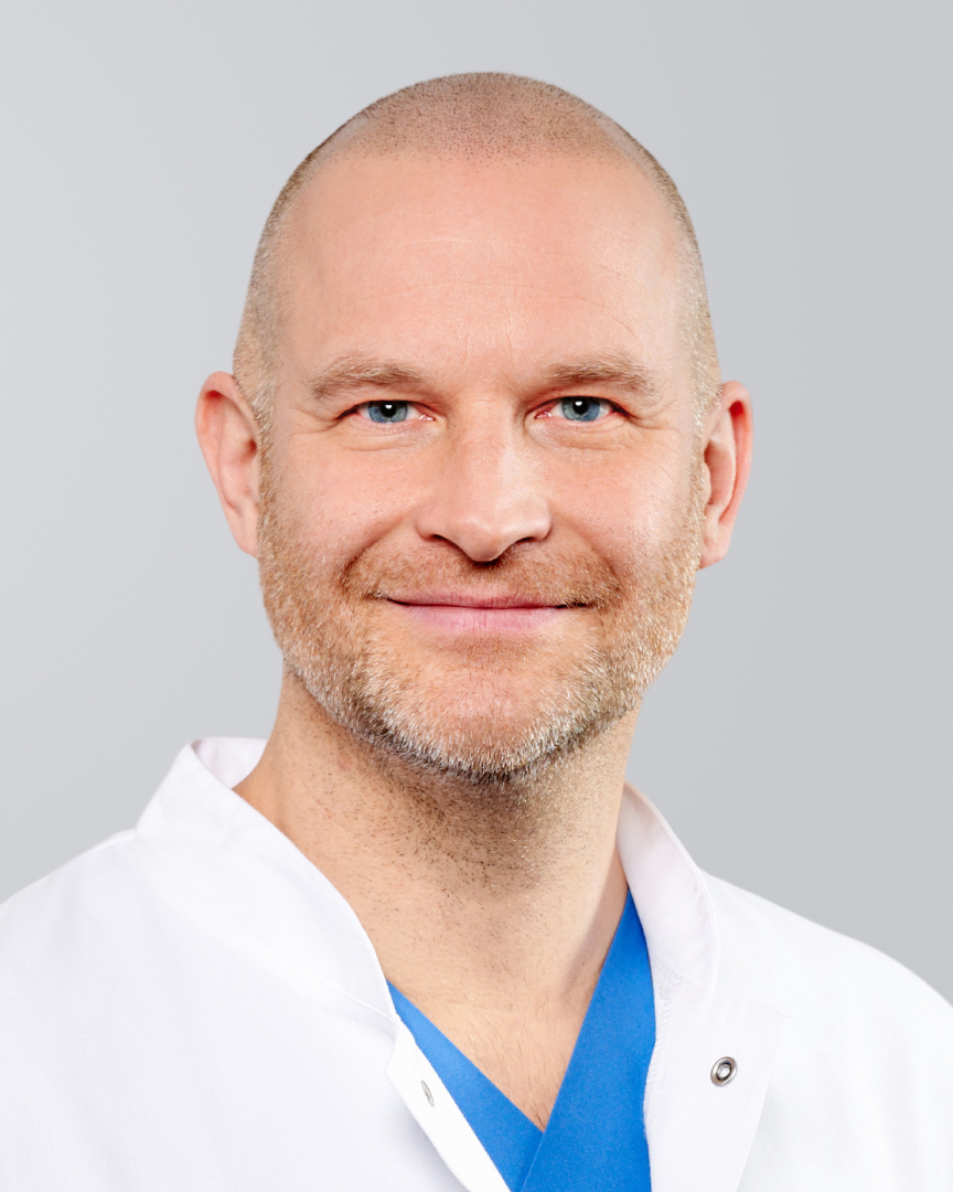 Lächelnder Mann mit Glatze und weißem Kittel vor grauem Hintergrund.