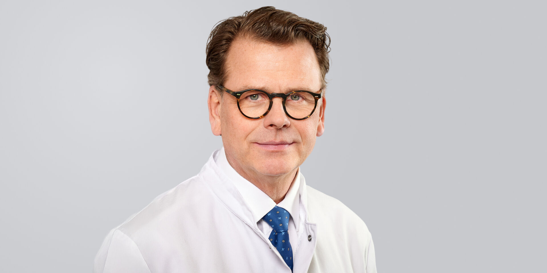 Lächelnder Mann mit Brille, Arztkittel und blauer Krawatte vor neutralem Hintergrund.