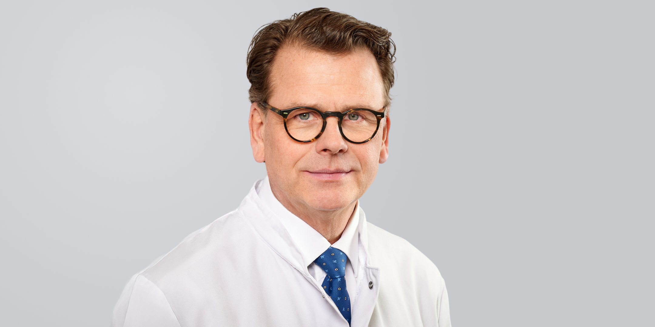 Lächelnder Mann mit Brille, Arztkittel und blauer Krawatte vor neutralem Hintergrund.