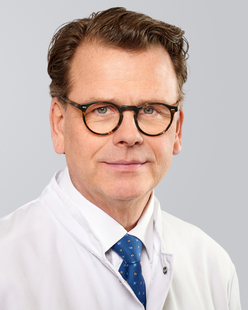 Lächelnder Mann mit Brille, Arztkittel und blauer Krawatte vor neutralem Hintergrund.