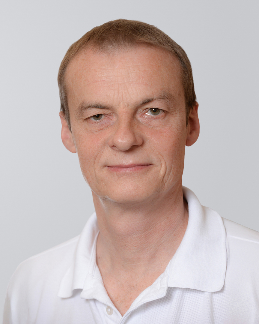 Porträt eines Mannes mit kurzem, blondem Haar, der in einem weißen Shirt vor einem hellen Hintergrund steht.