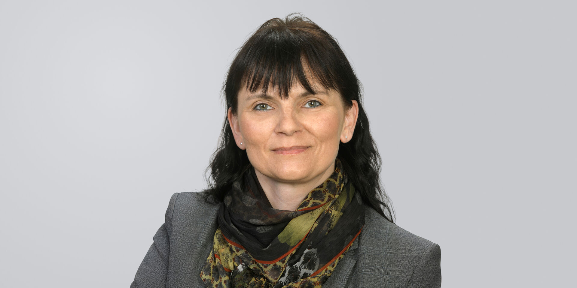 Porträt einer lächelnden Frau mit schwarzen Haaren und gemustertem Halstuch vor einem neutralen Hintergrund.
