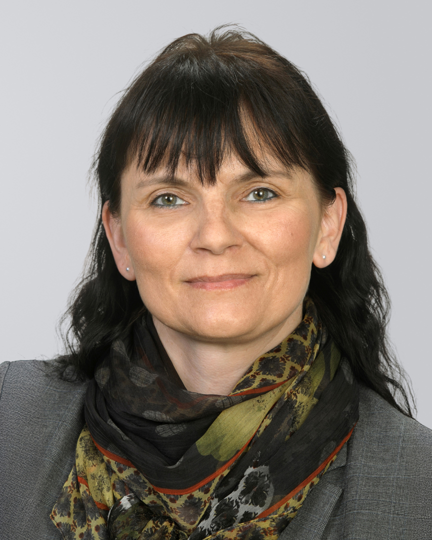 Porträt einer lächelnden Frau mit schwarzen Haaren und gemustertem Halstuch vor einem neutralen Hintergrund.