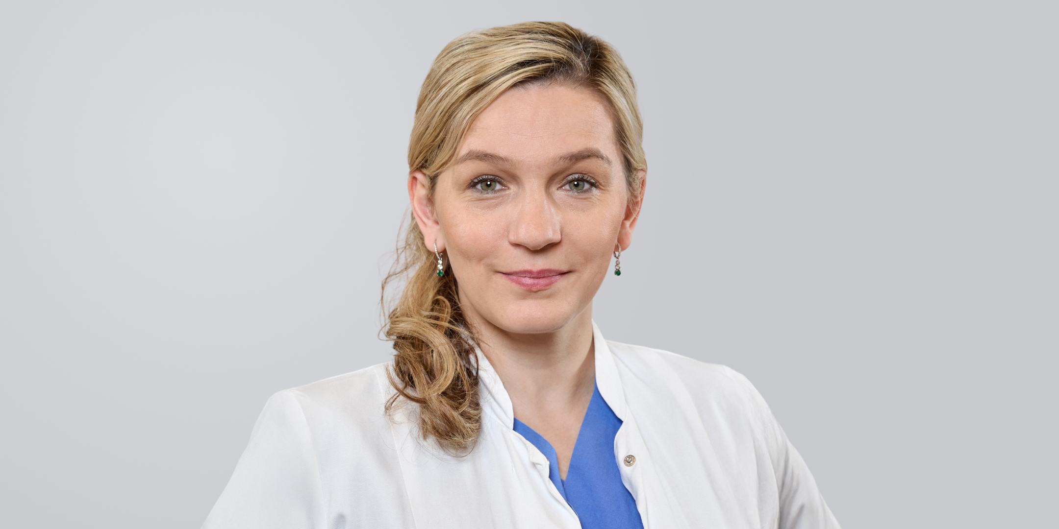 Frau mit blonden Haaren und weißem Kittel lächelt vor neutralem Hintergrund.
