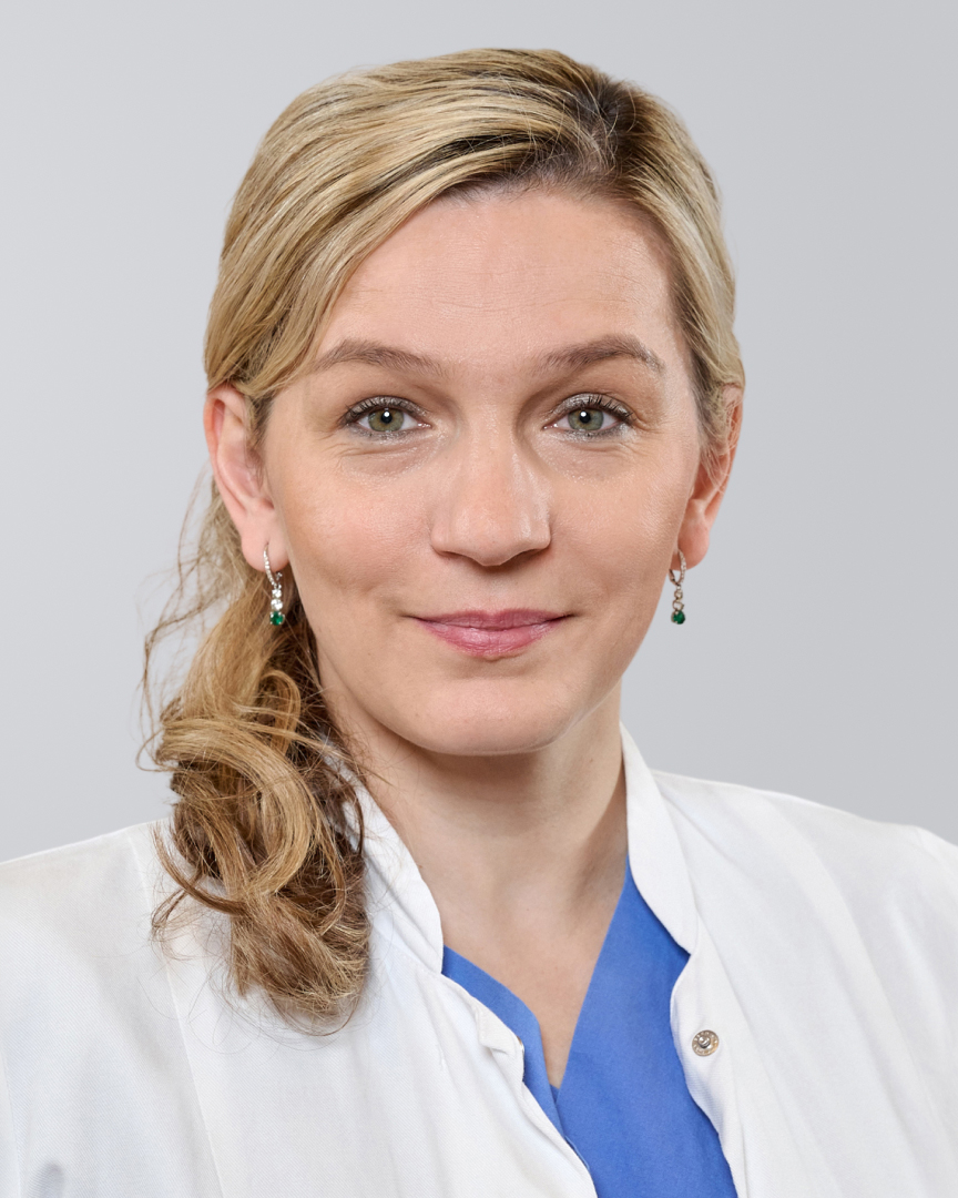 Frau mit blonden Haaren und weißem Kittel lächelt vor neutralem Hintergrund.