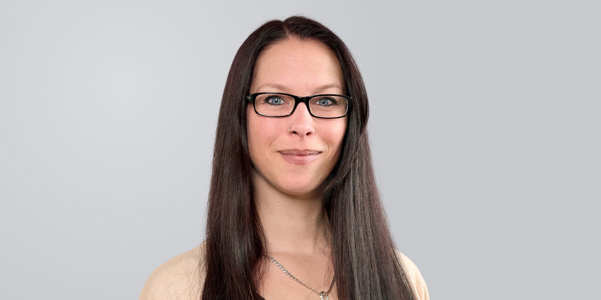Lächelnde Frau mit langen, glatten Haaren und Brille vor einem grauen Hintergrund.