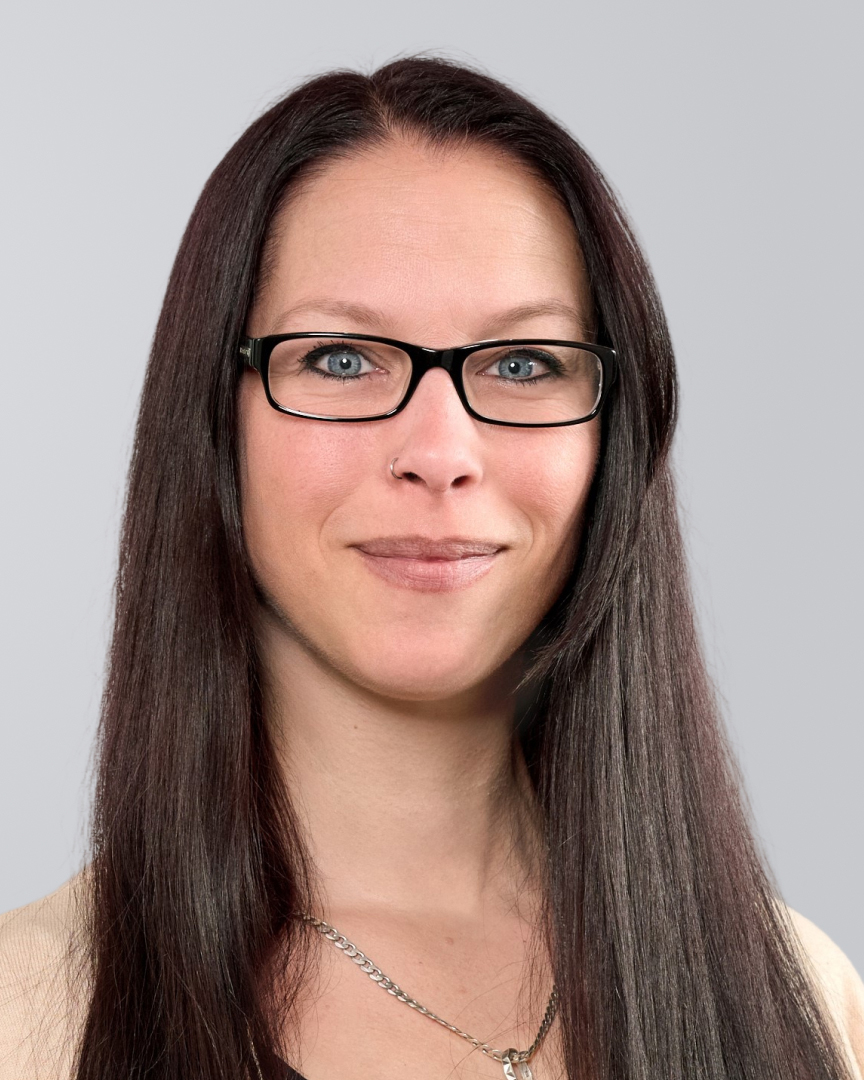 Lächelnde Frau mit langen, glatten Haaren und Brille vor einem grauen Hintergrund.