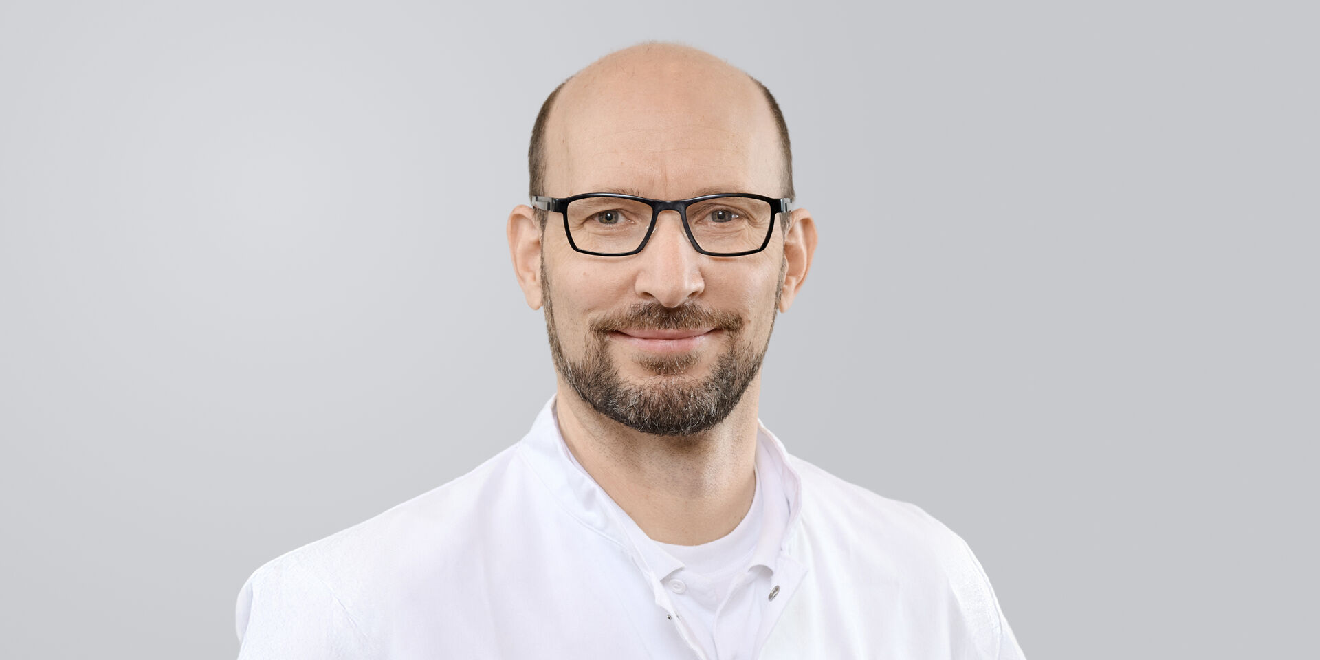 Mann mit Brille, Bart und weißem Kittel vor einem grauen Hintergrund.