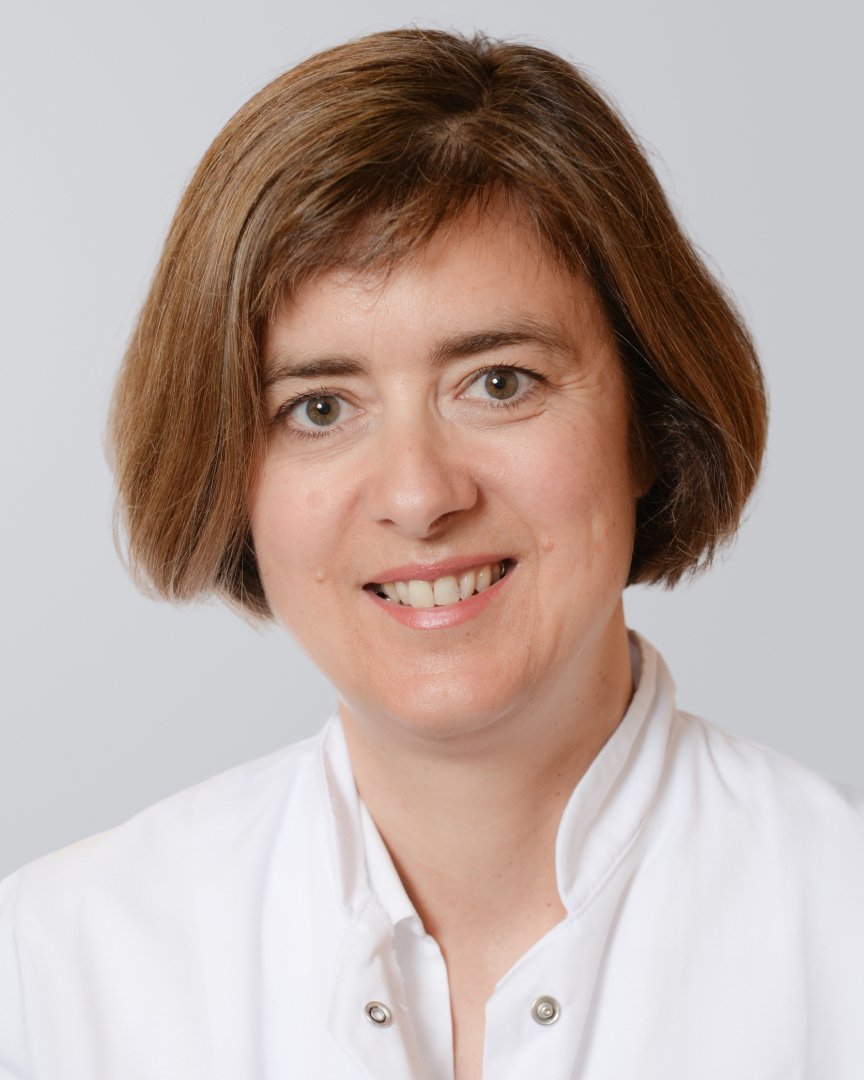 Lächelnde Frau mit kurzen, braunen Haaren in einem weißen Kittel vor einem hellen Hintergrund.