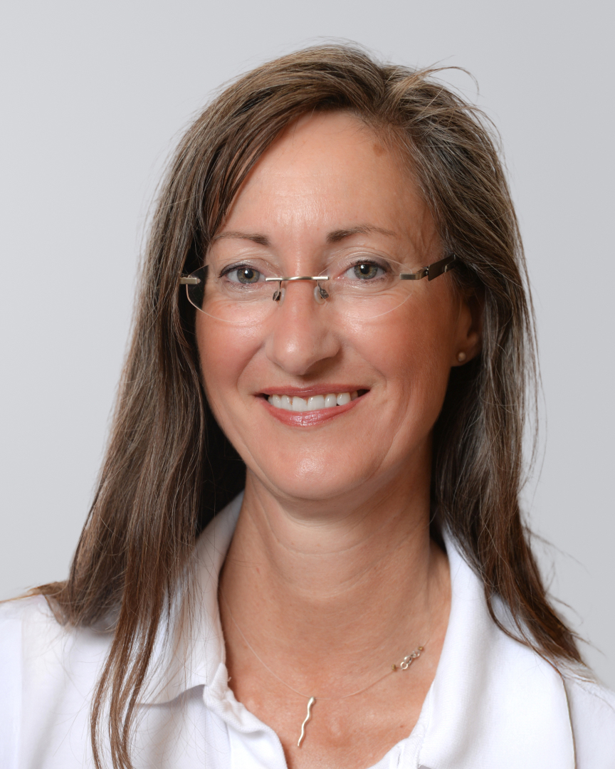Frau mit langen, braunen Haaren, Brille und einem weißen Oberteil vor neutralem Hintergrund.