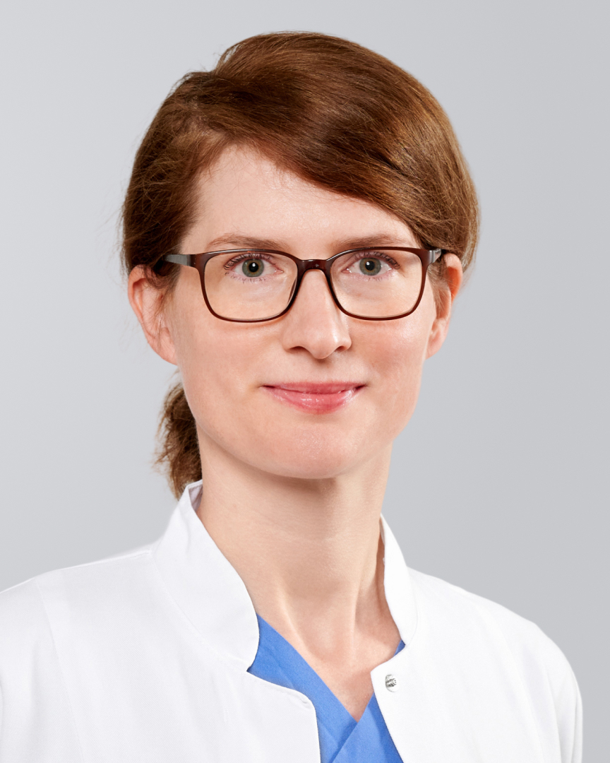 Junge Frau mit Brille, kurzen, braunen Haaren weißem Kittel vor neutralem Hintergrund.
