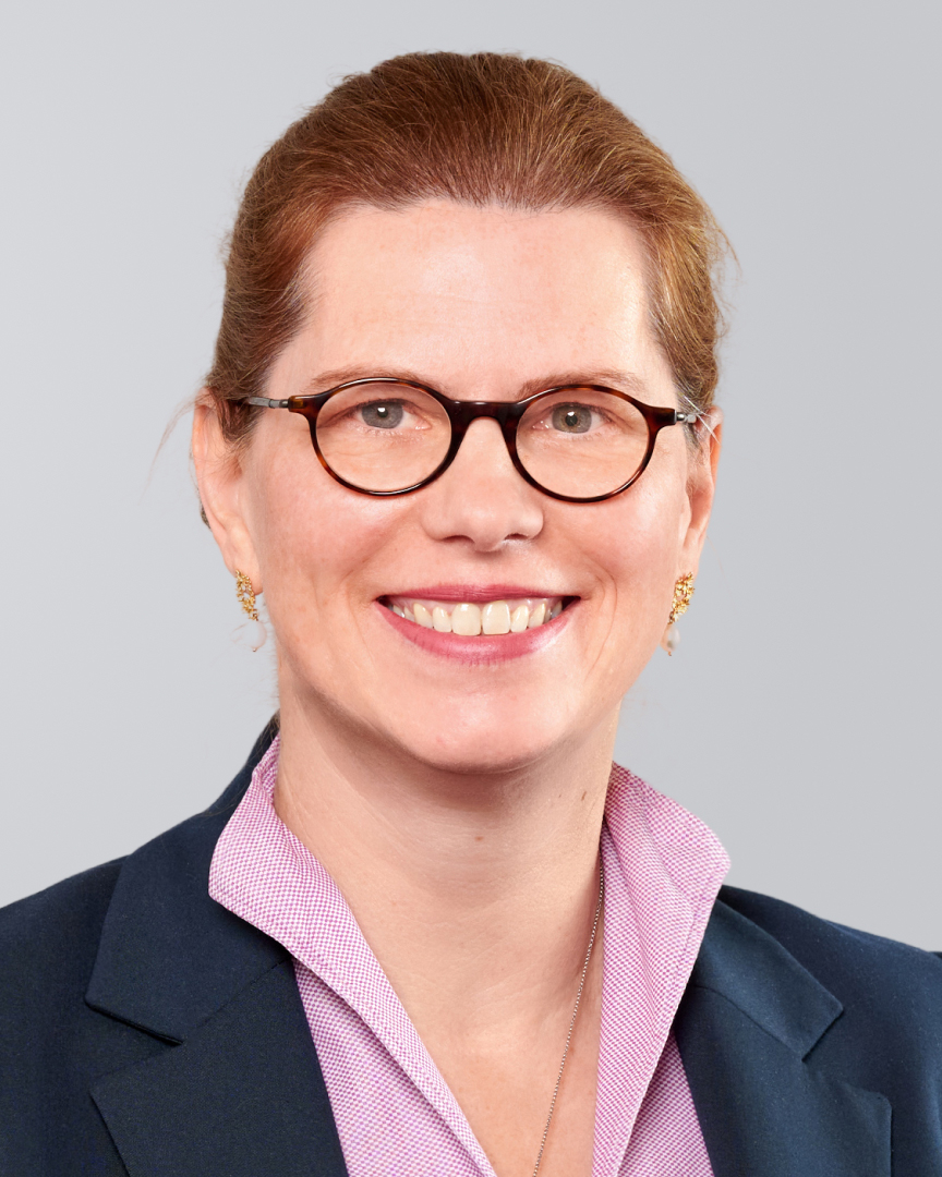 Frau mit Brille, rotem Haar und dunkelblauem Blazer vor einem neutralen Hintergrund.