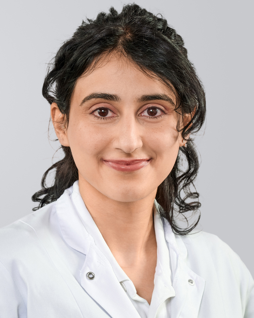 Frau mit dunklen, lockigen Haaren in einem weißen Kittel vor einem grauen Hintergrund.