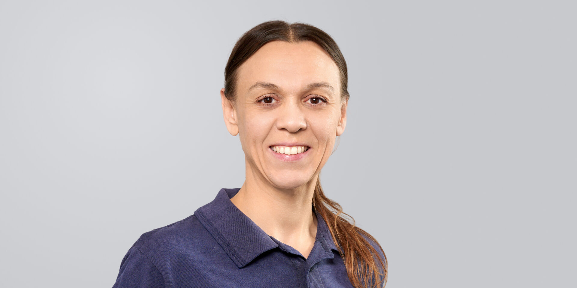 Porträt einer Frau mit langen, glatten Haaren und blauem Poloshirt vor einem neutralen Hintergrund.