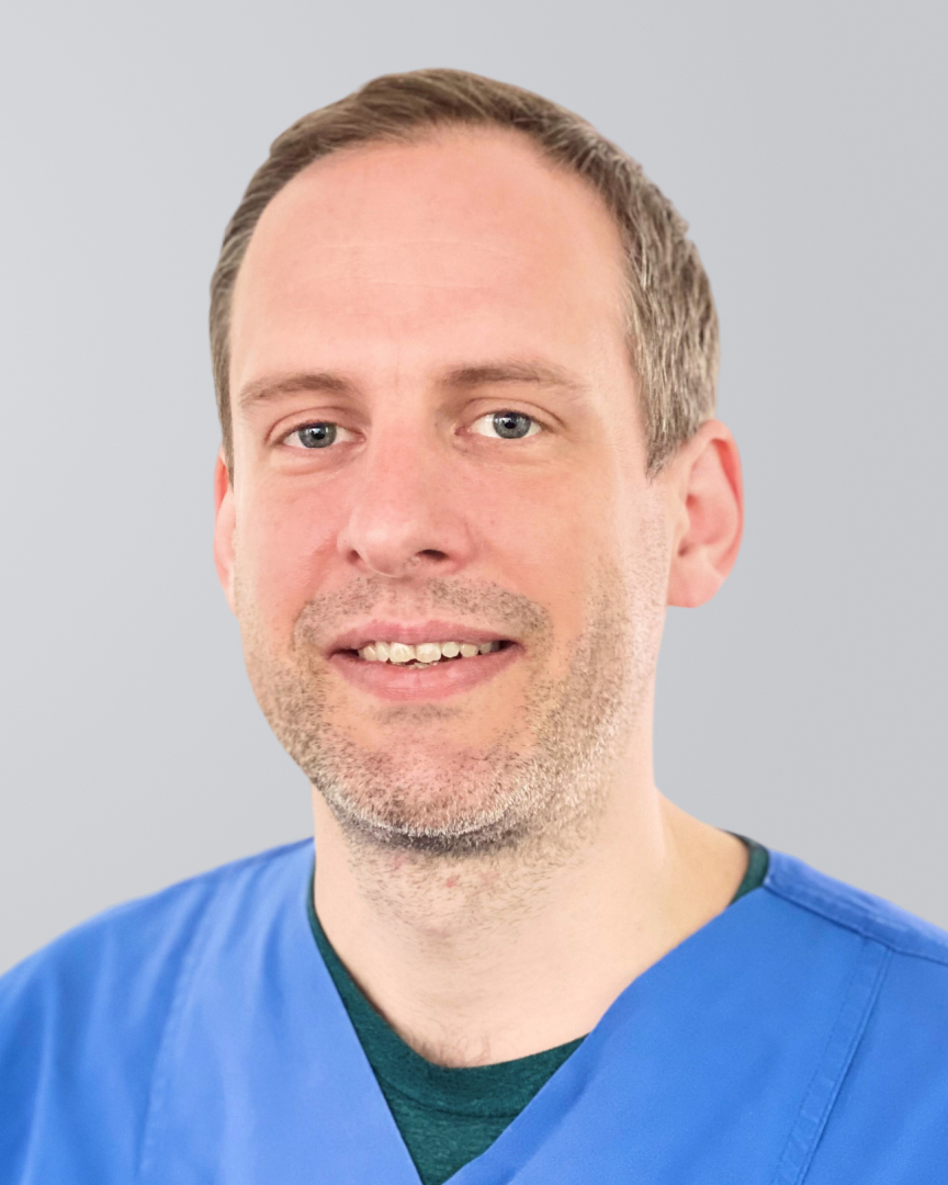 Lächelnder Mann mit kurzem Haar, Dreitagebart und blauer Arbeitskleidung vor neutralem Hintergrund.