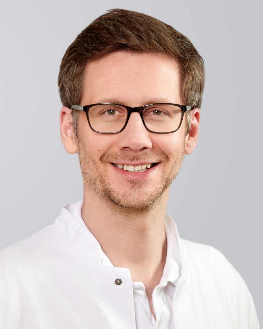 Lächelnder Mann mit Brille in weißem Kittel vor grauem Hintergrund.