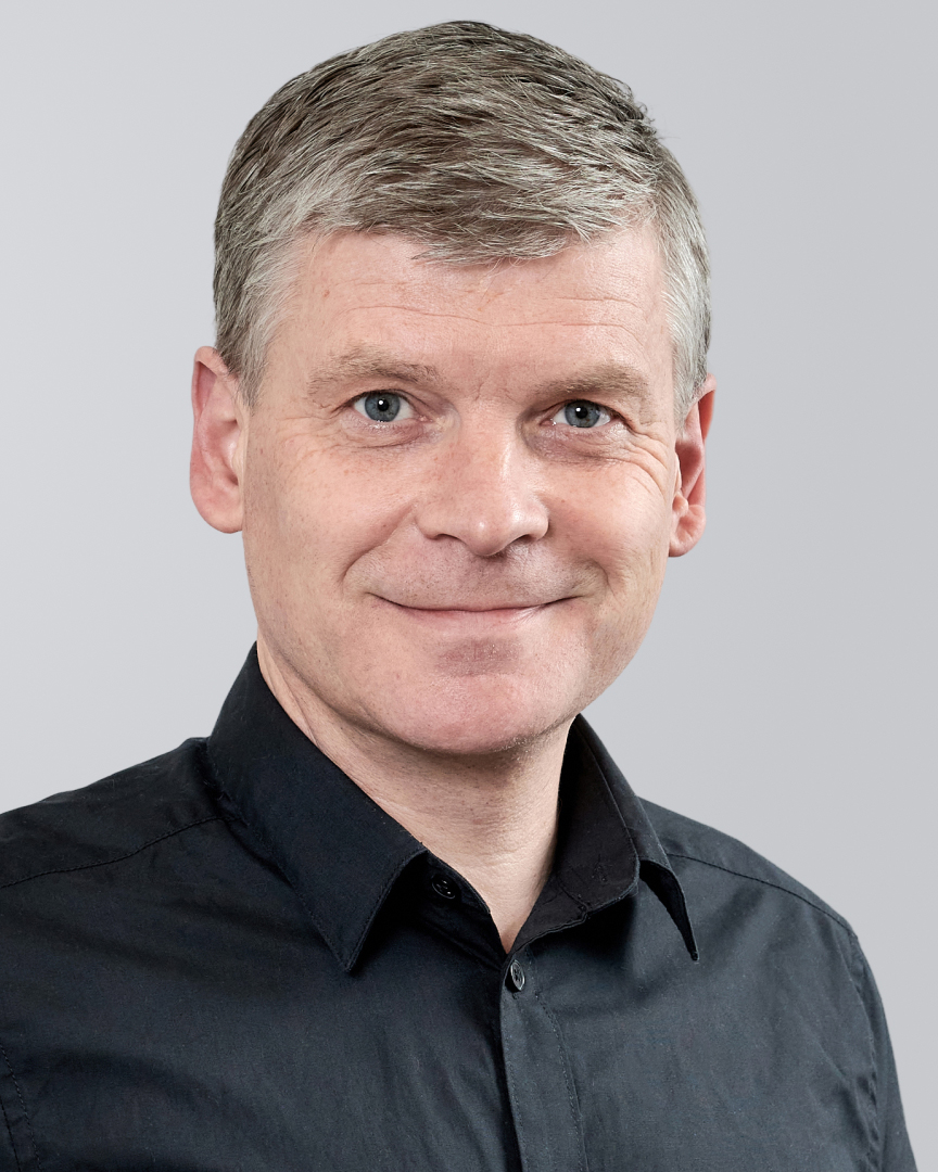 Mann mit kurzem, grauem Haar und schwarzem Hemd vor einem neutralen Hintergrund.