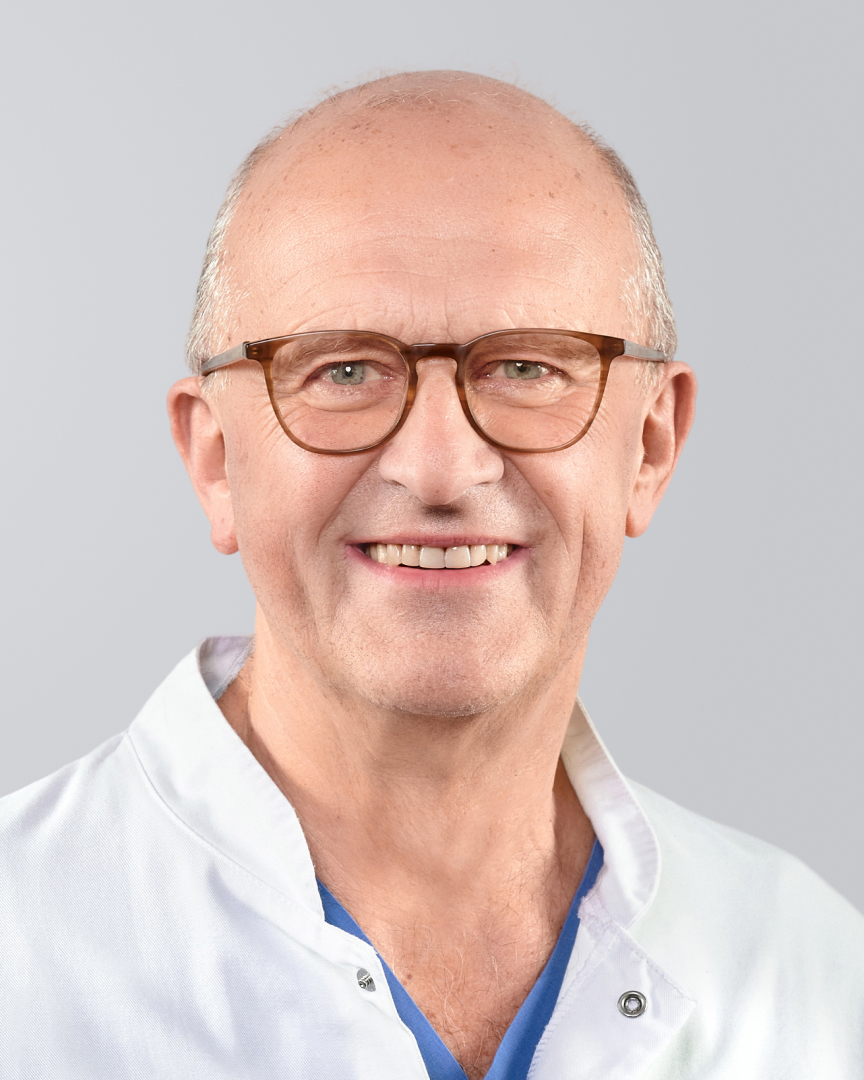 Lächelnder Arzt mit Brille, kurzen, grauen Haaren und weißem Kittel vor einem grauen Hintergrund.