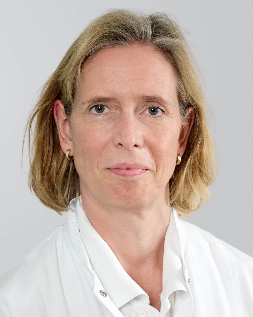 Frau mit blonden, schulterlangen Haaren und weißem Kittel vor einem grauen Hintergrund.