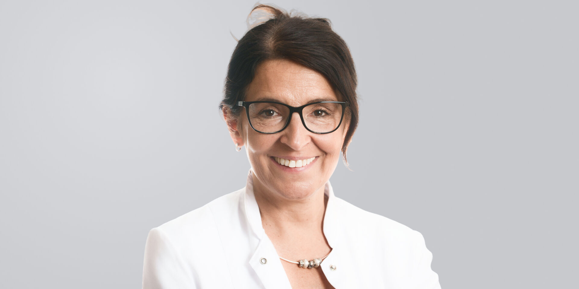 Lächelnde Frau dunklen Haaren, Brille und weißem Kittel vor einem grauen Hintergrund.