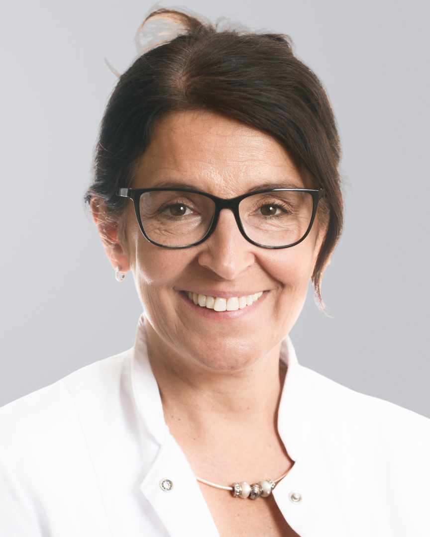 Lächelnde Frau dunklen Haaren, Brille und weißem Kittel vor einem grauen Hintergrund.