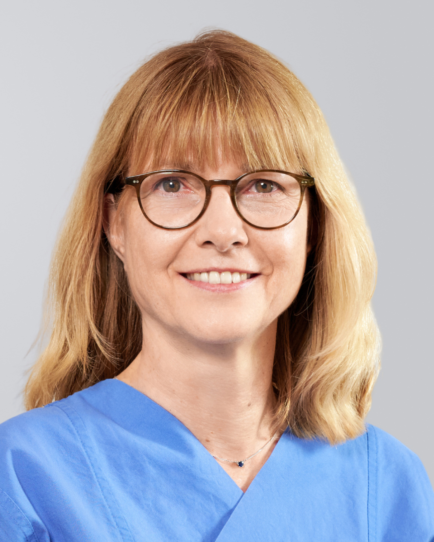 Lächelnde Frau mit blonden Haaren, Brille und blauem medizinisches Outfit vor neutralem Hintergrund.