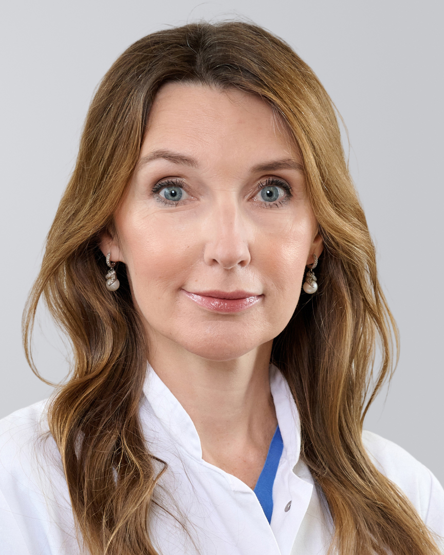 Frau mit weißem Kittel und langen, braunen Haaren vor einem grauen Hintergrund.
